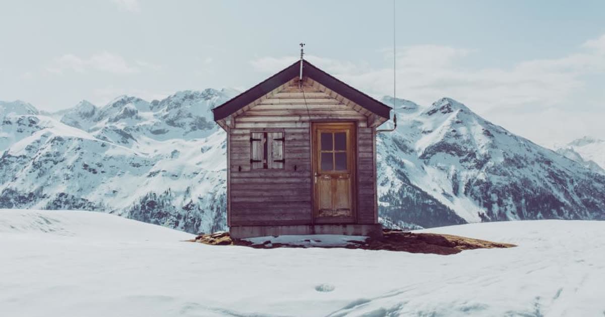 A small wooden cabin in deep snow with mountains behind it